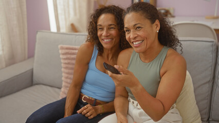 Obraz premium Women laughing together at home on a couch holding a remote in a cozy living room with natural light, showcasing friendship and family bonds.