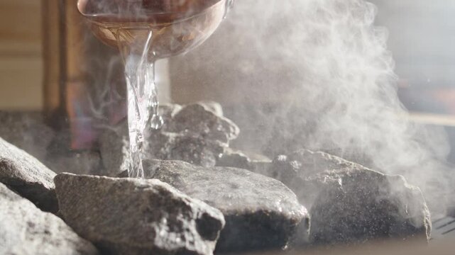Water sizzling and steaming as copper ladle pours it over sauna rocks, closeup
