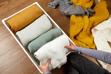 The hands of a woman neatly folding knitted clothes into a metal laundry basket. The concept of restoring order, cleaning, cluttering, organizing space.