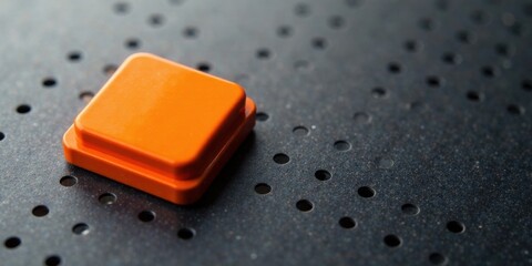 A vibrant orange square block rests on a dark perforated surface, subtly highlighting a contrast in texture and color.