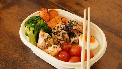 A healthy bento box with grilled salmon, steamed broccoli, cherry tomatoes, roasted sweet potatoes, and a boiled egg, served with chopsticks on a rustic wooden table.