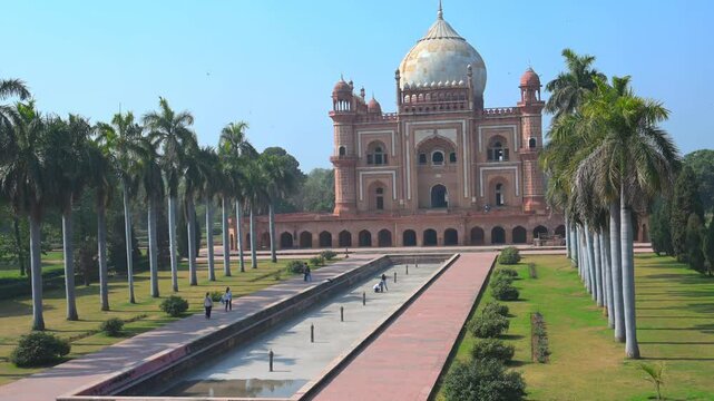 The Safdarjung Tomb in Delhi, an exquisite Mughal-era structure, features an iconic dome, intricate red sandstone facade, lush gardens, a long symmetrical pathway, and towering palm trees under a brig