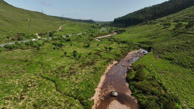 Barnesmore Gap, County Donegal, Ireland, June 2023. Drone pushes forwards ascending above a river, slowly approaching a road as cars zoom past below a dense forested valley on a warm summer day.