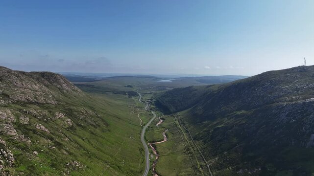 Barnesmore Gap, County Donegal, Ireland, June 2023. Drone pulls back between steep, exposed rock cliffs on the mountains as a river meanders alongside a winding N15 road on a summer day.