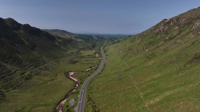 Barnesmore Gap, County Donegal, Ireland, June 2023. Drone push in follows meandering road through the lowlands next to river with sandy banks on a blue sky day, cars drive quickly to town