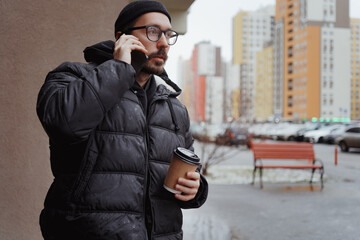 A young man in casual style walks through the snowy city on a winter day, chatting on his phone and sipping coffee. The lifestyle photo captures his relaxed, modern vibe and cheerful energy