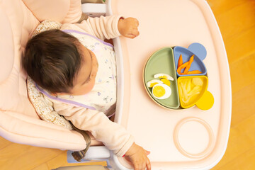 baby sitting in feeding highchair with pink tray with colorful plate of healthy food cut into pieces. Top view Baby led weaning method