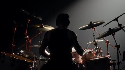 Cinematic shot with silhouette of professional rock musician playing drum kit. Back view of young adult brawny man sitting alone in rehearsal room and rehearsing, backlit silhouette in darkness