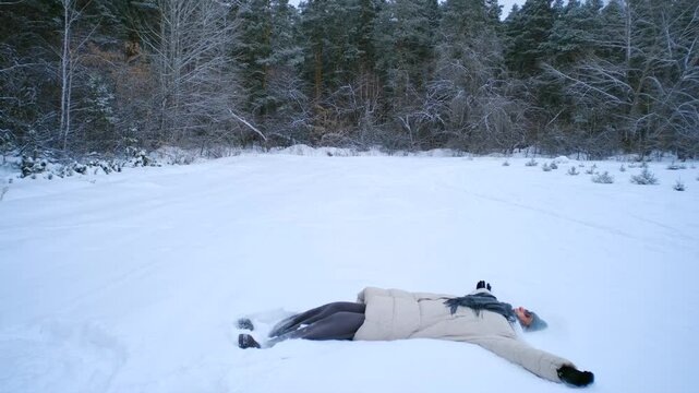 Young woman falling backward in snowy forest, creating snow angel with joyful expression, surrounded by serene, snow-covered trees during peaceful winter landscape moment