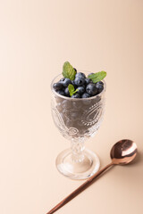 Cut glass cup filled with blueberries with mint next to a copper spoon on neutral background. Minimalist styling. Copy Space. Healthy food concept, freshness and refined style