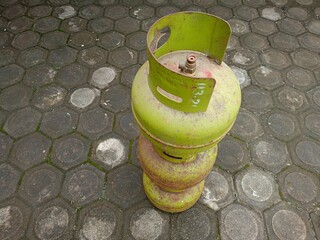 A rusty green 3 kg LPG gas cylinder stands on a paving block floor. The photo was taken from a top...