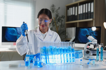 Female scientist wearing lab coat and protective glasses using micropipette dropping blue liquid...