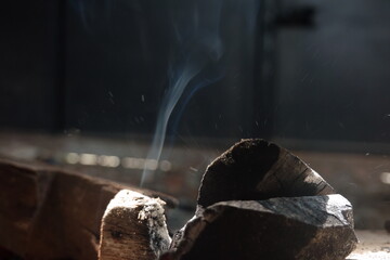 Close-up view of smoldering wood emitting smoke in a dimly lit setting, evoking warmth, calm, and...
