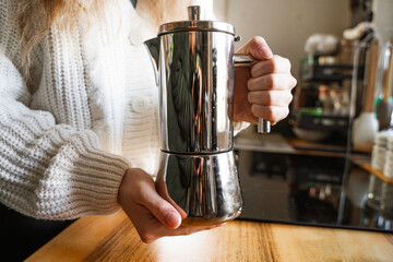 A woman holding a stainless steel moka pot for making hot fresh coffee drinks at home in the kitchen. Close-up of a metal coffee pot in a girl's hands. Equipment for making coffee. Coffee brewing.