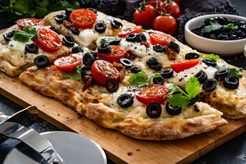 Pinsa Romana with burrata cheese, black olives and cherry tomatoes on black table	