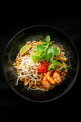 A vibrant plate of Pad Thai featuring shrimp, bean sprouts, fresh lime, peanuts, and cilantro, beautifully arranged on a dark background, highlighting its delicious textures and colors.
