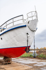 White and red ship raised on blocks for maintenance at dry dock near river, with cloudy sky and...