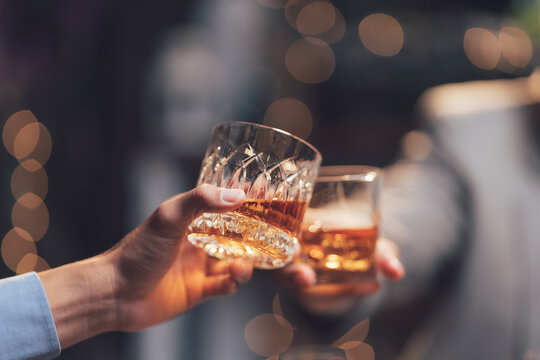Cheers to Connection: Two hands clink glasses of amber liquid, a toast to friendship, celebration, or a shared moment of enjoyment, bathed in the soft glow of warm lights.