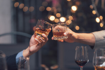 Toasting the Good Times: Close-up of two hands clinking glasses of amber liquid, with warm, blurred lights creating a celebratory ambiance.