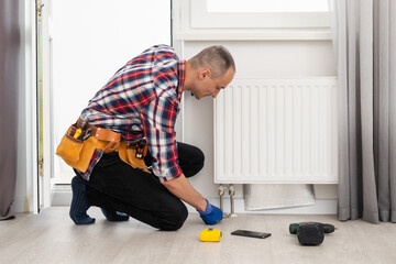A plumber in a blue jumpsuit is fixing a radiator. With an adjustable wrench, he tightens the tap....