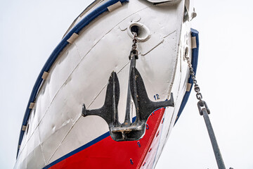 Ship's bow with a red and white hull, flanked by two large anchors on chains close-up. Vessel is...