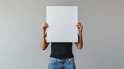 person holding a blank white poster in front of their face. The person is wearing blue jeans and a black t-shirt.