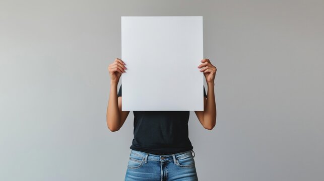 person holding a blank white poster in front of their face. The person is wearing blue jeans and a black t-shirt.