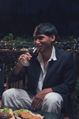 A Toast to the Evening: A young man dressed in a stylish blazer savors a glass of red wine while enjoying a delectable meal in the tranquil ambiance of an outdoor dining setting.