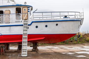 White and red ship raised on blocks for maintenance at dry dock near river, with cloudy sky and...