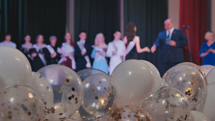 White and transparent balloons with gold and silver confetti fill the foreground, celebrating a high school graduation ceremony on stage
