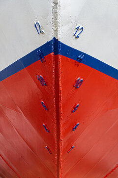 Ship's bow with red, white, and blue paint, featuring blue draft markings on weathered metal and welded seams detailed close-up