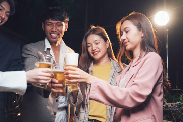 Nightlife Cheers: Young adults, a diverse group of friends, raise their beer glasses in a celebratory toast under the warm glow of outdoor night lighting.  A moment of camaraderie and festive joy. 