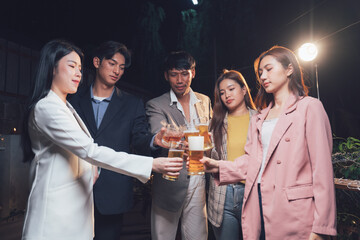 Nighttime Cheers: A group of young adults, dressed in stylish attire, raise their drinks in a celebratory toast under the warm glow of outdoor lighting.