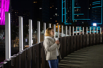 A woman holding a coffee cup on a well-lit bridge at night, surrounded by urban architecture and...