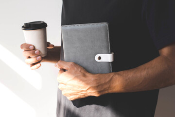A man in a black t-shirt holding a stylish gray notebook with a white clasp and a takeaway coffee cup in natural sunlight, symbolizing productivity and modern lifestyle