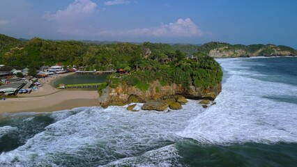 Aerial Drone view of the beach with white sand, coral rocks, hills with trees, waves from the ocean which is a tourist destination known as Drini Beach Gunung Kidul Yogyakarta