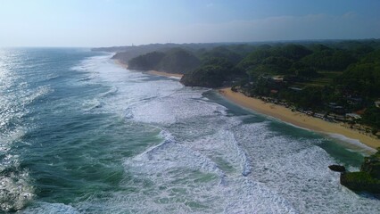 Aerial Drone view of the beach with white sand, coral rocks, hills with trees, waves from the ocean which is a tourist destination known as Drini Beach Gunung Kidul Yogyakarta