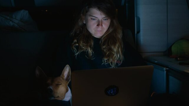 Digital nomad woman work remotely on project on her laptop inside a camper van at night. Blue screen lit face. Working remotely as a creative. Living off grid in a converted camper. Travel with dog