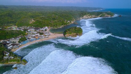 Aerial Drone view of the beach with white sand, coral rocks, hills with trees, waves from the ocean which is a tourist destination known as Drini Beach Gunung Kidul Yogyakarta
