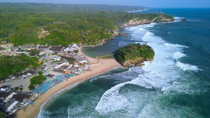 Aerial Drone view of the beach with white sand, coral rocks, hills with trees, waves from the ocean which is a tourist destination known as Drini Beach Gunung Kidul Yogyakarta