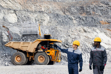 Inspecting open mine, excavator loading sand into truck. Caucasian male and african man wearing...