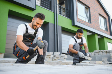 A group of multiracial workers lay paving stones on a sidewalk near a house