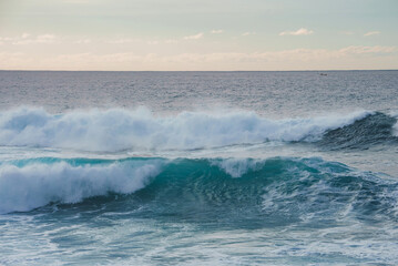 Large turquoise waves roll in the foreground, a small boat is visible on the horizon, and pastel toned clouds fill the sky in a coastal Hawaii scene.