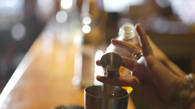 A person is pouring a drink into a glass. The drink is a mixture of alcohol and other ingredients. The person is holding a shaker and a glass. The scene is set in a bar or a similar establishment
