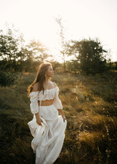 Woman in a flowing white lace dress walks through a sunlit field, in golden hour light.