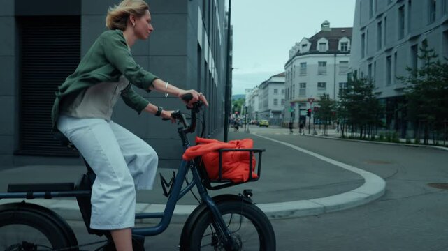 Cute adorable and stylish young woman pedal through town on cargo electric bike. Student or creative office worker cycle in business district on a warm summer morning