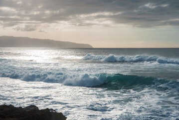 Serene ocean scene with waves hitting rocky shores, a mountainous Hawaiian coastline in the distance, and sunlight reflecting under cloudy skies.