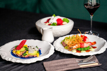 An elegant dining setup featuring two gourmet dishes: one with vibrant ingredients and a beautifully presented prawn dish, accompanied by a glass of red wine and a dessert in the background.