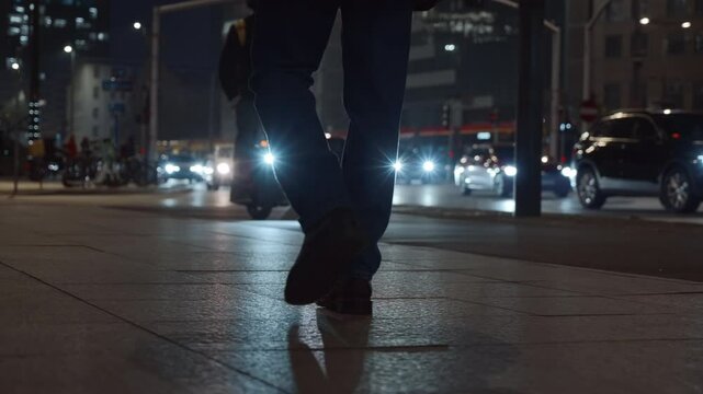 Rear side view male feet steps walking street at night. Man wearing dark leather boots walks pedestrian crossing in city backlit with car headlights. Low angle people legs going sidewalk, cold weather