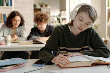 Portrait of young woman wearing headphones while sitting at desk in college library focused on...